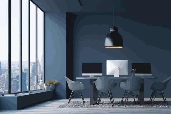 Coin bureau fonctionnel moderne avec trois écrans sur une table partagée, chaises design grises, suspension industrielle noire et grande baie vitrée sur vue urbaine.