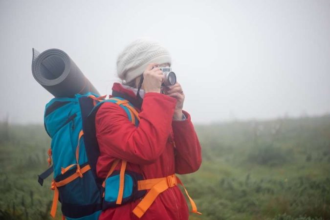 Une randonneuse avec un grand sac à dos et un bonnet prend une photo avec un appareil argentique dans un paysage montagneux brumeux, immortalisant ses randonnées en voyage.