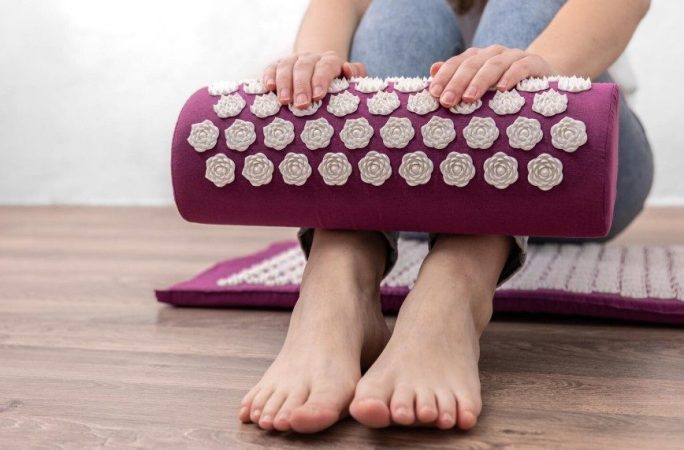Personne assise tenant un coussin d'acupression violet au-dessus d'un tapis de massage champ de fleurs utilisation dos.