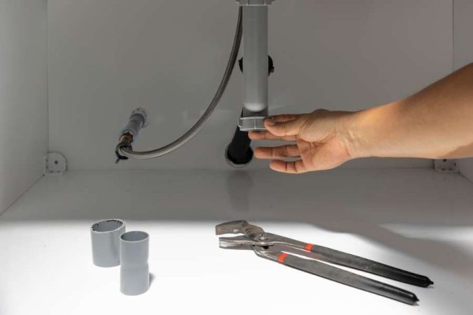 Une main resserre l'écrou en plastique d'un siphon de lavabo ou d'évier à l'intérieur d'un meuble blanc, avec une pince multiprise posée au premier plan.