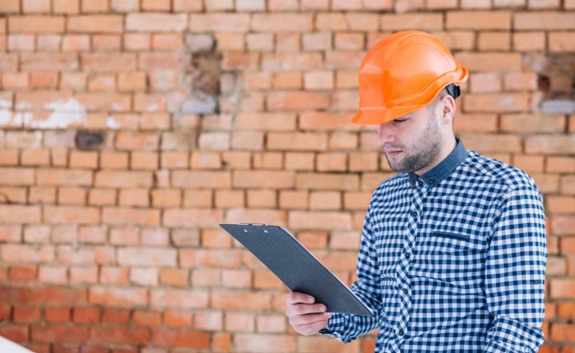 Un expert en bâtiment portant un casque orange et une chemise à carreaux examine des documents sur un presse-papiers devant un mur en briques anciennes. L'inspection est cruciale pour qui souhaite acheter une maison ancienne.