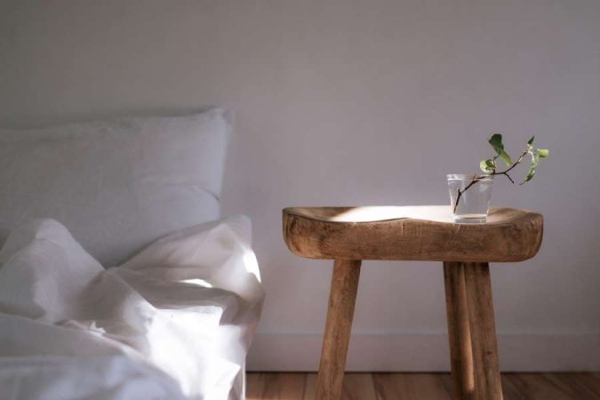 Une table d'appoint rustique en bois massif est placée dans une chambre lumineuse à côté d'un lit aux draps blancs.